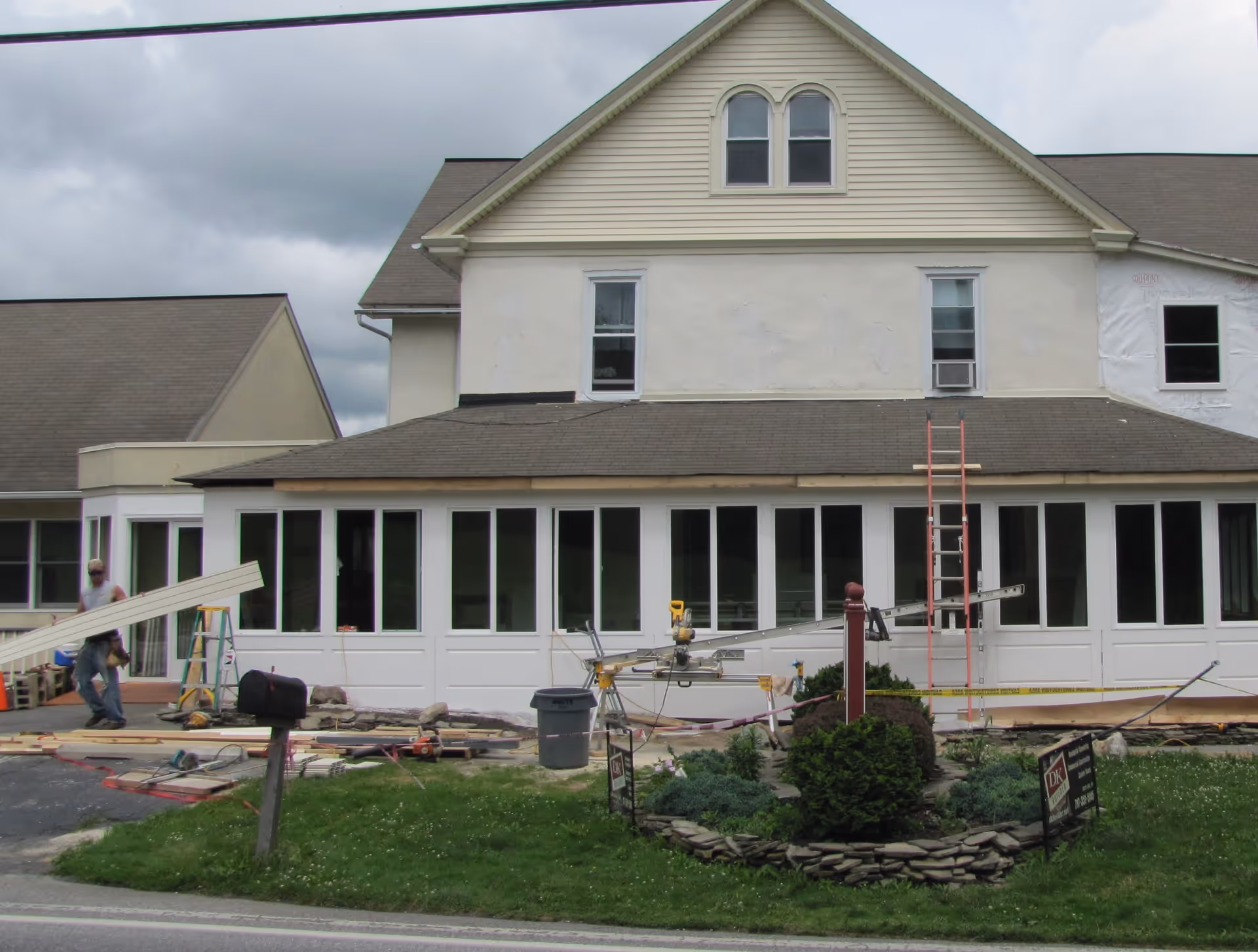Exterior view of a building under renovation with construction materials and equipment in front. A worker is carrying wooden planks near the left side. The building has multiple windows and a gabled roof, with a small landscaped area featuring bushes and a mailbox in the foreground.