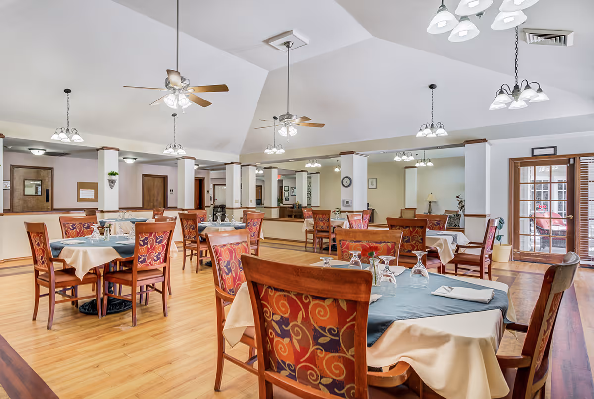 Spacious, well-lit dining room with tables set with linens and glassware, patterned wooden chairs, ceiling fans and pendant lights.