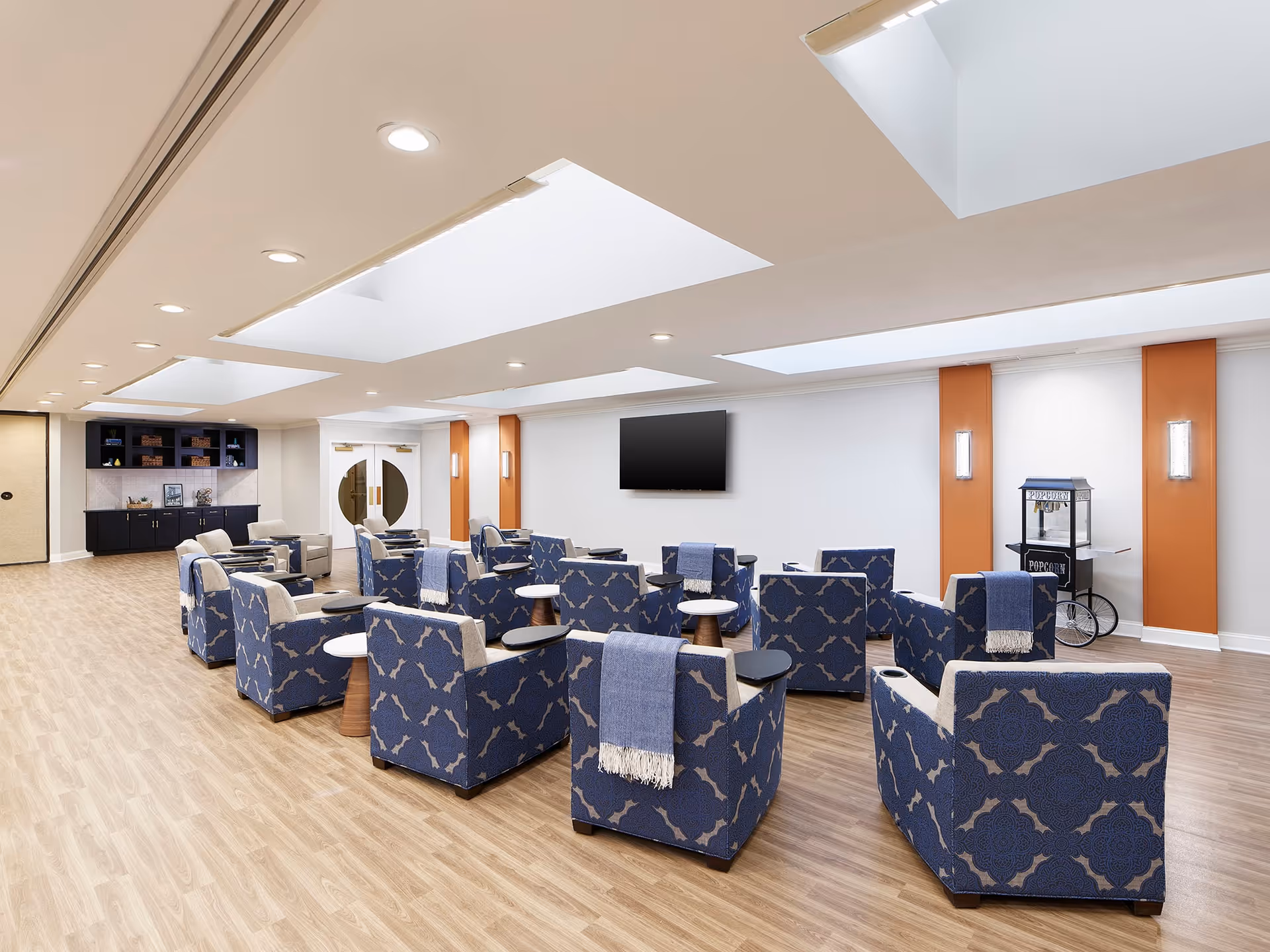 A spacious senior living common area with patterned blue armchairs arranged in rows facing a wall-mounted flat screen TV. The room has light wood flooring, white walls with orange accent panels, and skylights in the ceiling. There is a popcorn machine on the right side and a black cabinet with shelves on the left side near double doors with circular windows.