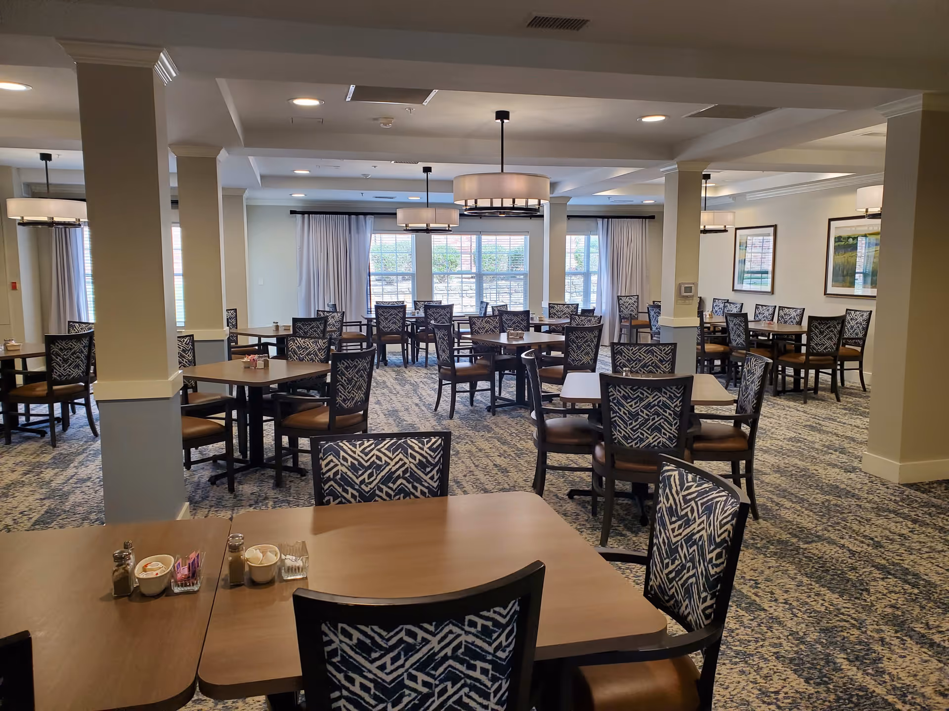 A spacious dining room with multiple wooden tables and chairs featuring patterned upholstery. The room has carpeted floors, several columns, large windows with curtains letting in natural light, and modern ceiling light fixtures. The walls are decorated with framed artwork.