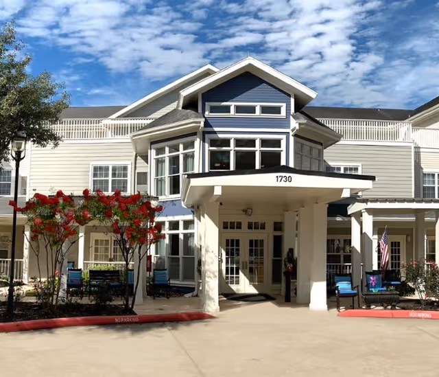 Front exterior view of Sundale Senior Living of The Woodlands building with a covered entrance, large windows, and a clear blue sky with scattered clouds. There are red flowering trees and outdoor seating near the entrance.