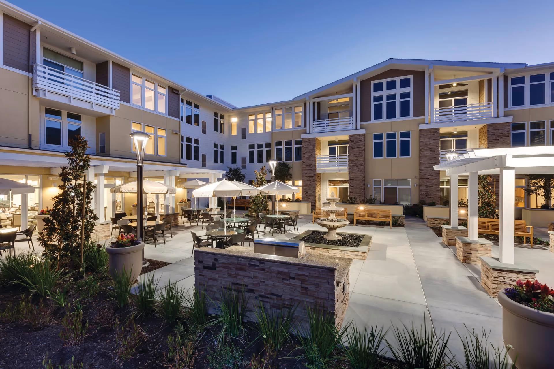 Outdoor courtyard area of a senior living facility at dusk with tables, chairs, and umbrellas arranged around a central fountain. The building surrounding the courtyard has multiple windows and balconies, with warm lighting illuminating the scene. There are benches and landscaped plants along the pathways.