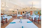 A spacious dining room with multiple tables covered in white tablecloths, each set with blue napkins and a small flower centerpiece. The room features wooden chairs, chandeliers hanging from the ceiling, and large windows allowing natural light to fill the space.