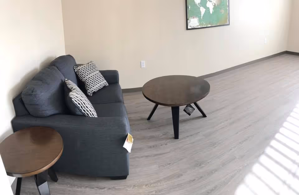 A small living area with a dark gray sofa adorned with two patterned pillows, a round wooden coffee table, and a matching side table. The room has light-colored wooden flooring and beige walls, with a framed world map hanging on the wall.