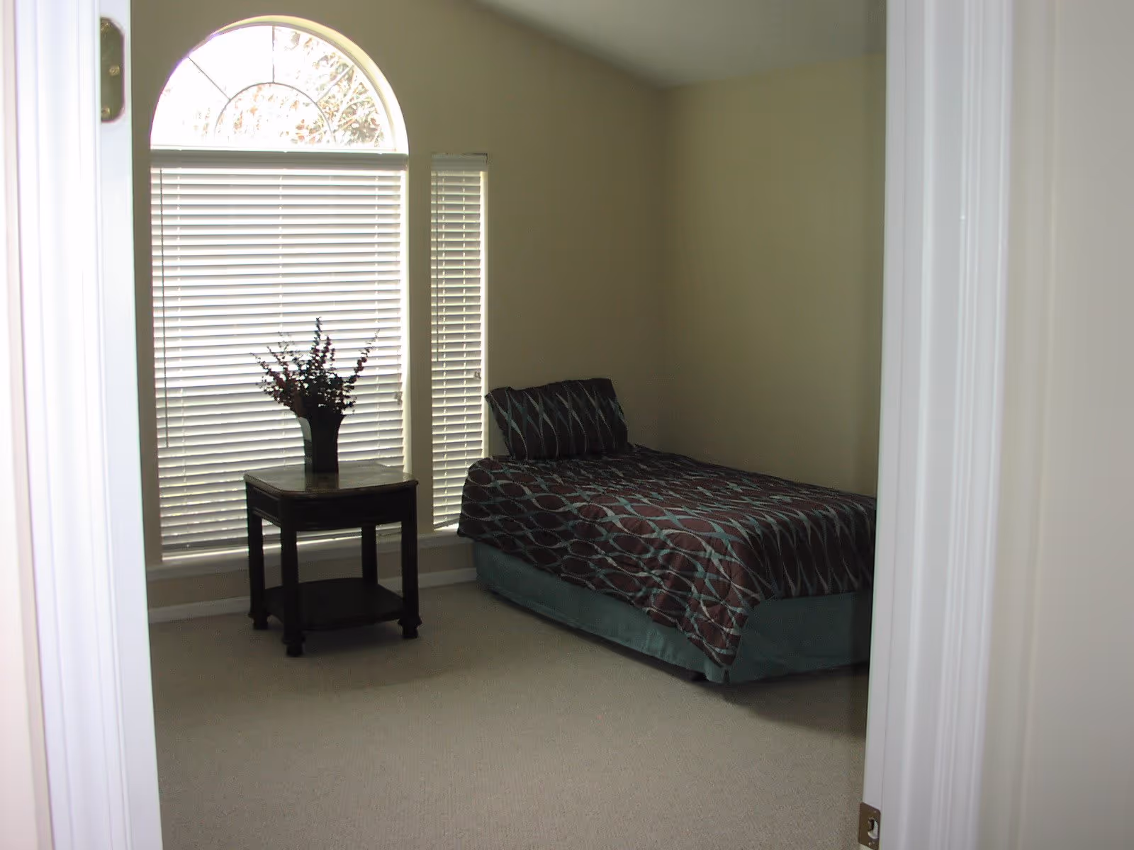 A simple bedroom with a single bed covered in a patterned bedspread and a matching pillow. Next to the bed is a small wooden side table with a vase of flowers. The room has beige walls and carpet, and a large window with white blinds and an arched top letting in natural light.