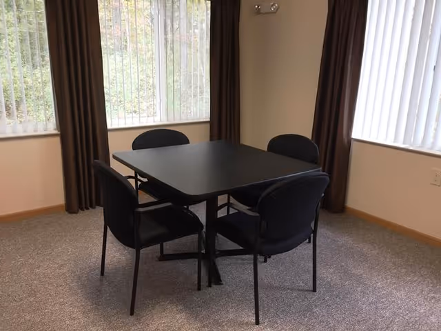 A small square black table with four black chairs around it in a carpeted room with two large windows covered by vertical blinds and brown curtains.