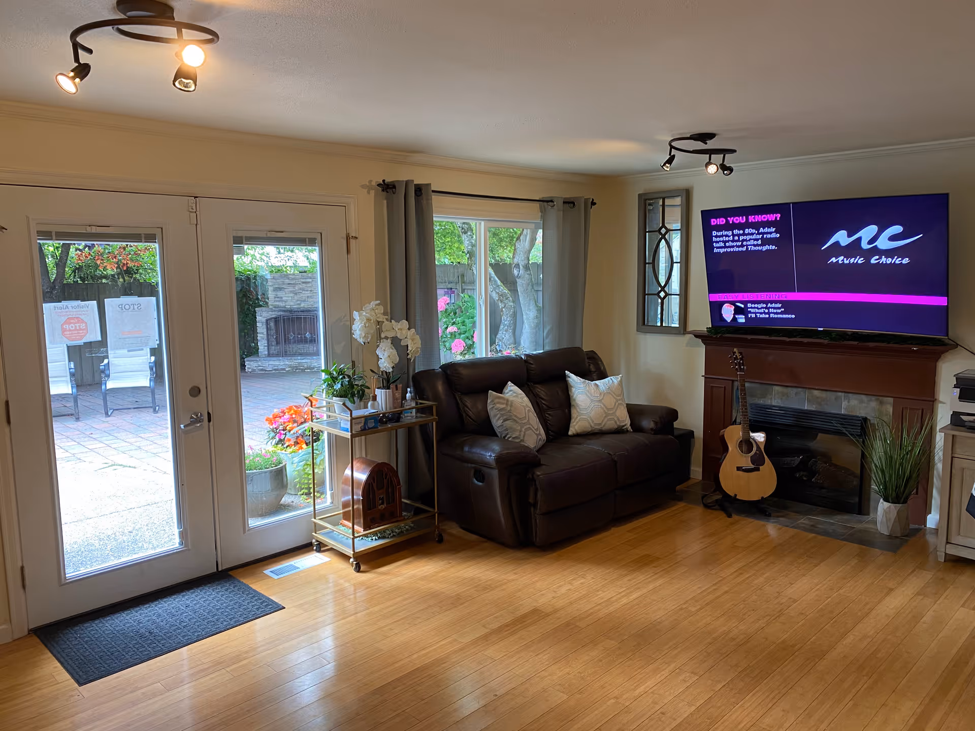 A cozy living room with a brown leather loveseat adorned with two patterned pillows, a small gold and glass side table holding plants and a decorative item, a mounted flat-screen TV displaying Music Choice, a wooden fireplace with a guitar resting against it, and a large window and glass doors leading to an outdoor patio with chairs and a fireplace.