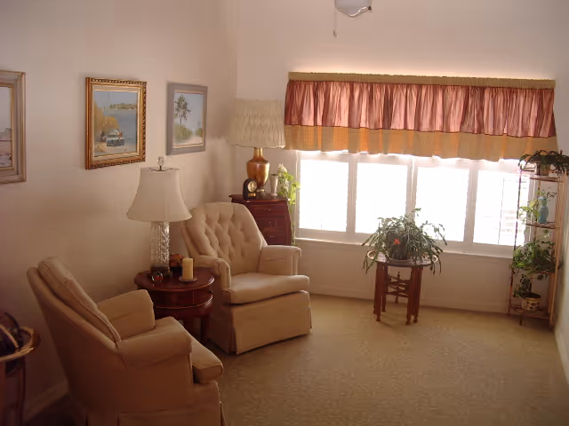 A cozy living room with two beige upholstered armchairs, a round wooden side table with a lamp and candle, a wooden chest with a lamp and clock, several framed paintings on the wall, a large window with a pink and gold valance, and multiple potted plants on stands near the window.