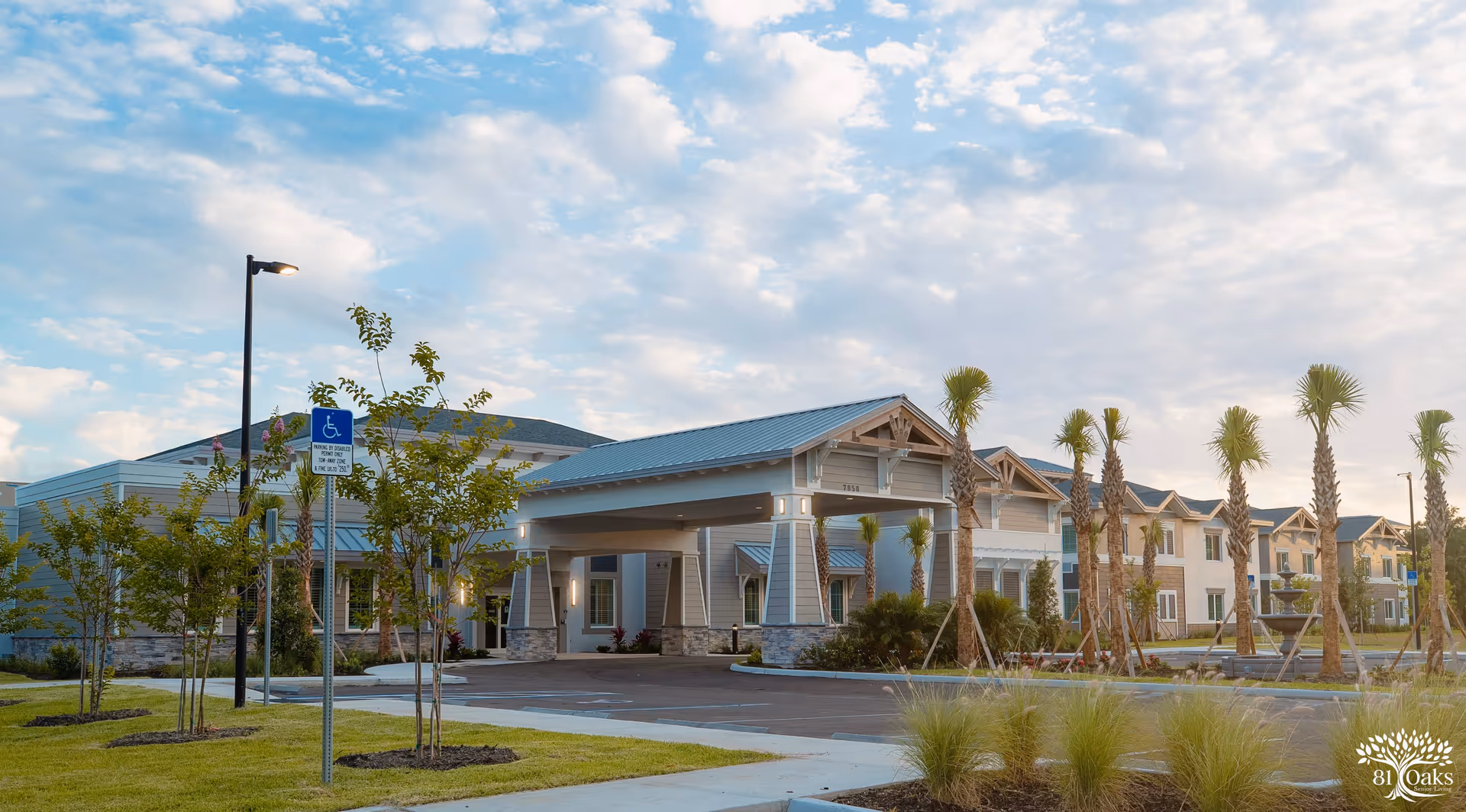 Exterior front view of 81 Oaks Senior Living with a covered entrance, palm trees, and parking under a partly cloudy sky.