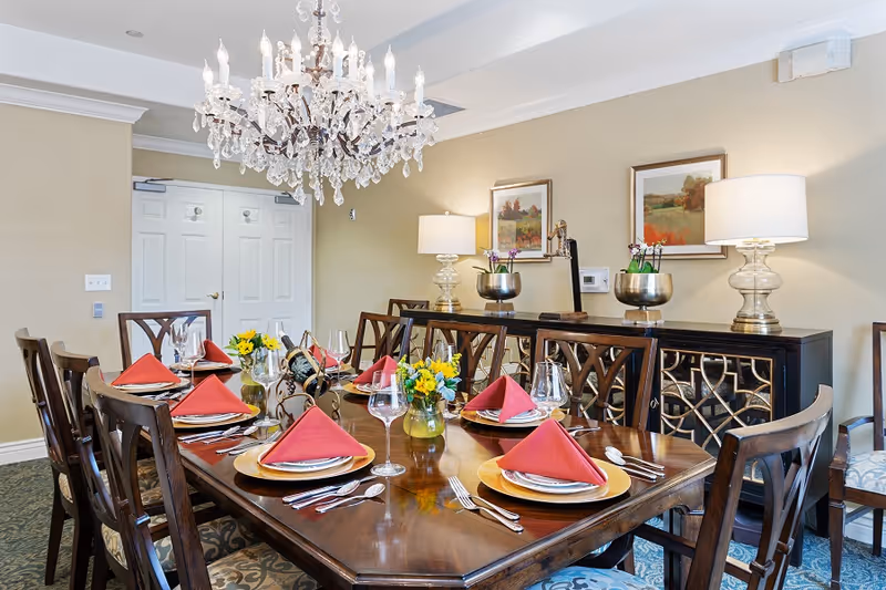Elegantly set dining room with a long wooden table, red napkins, floral centerpieces, a crystal chandelier, and a sideboard with lamps and artwork.