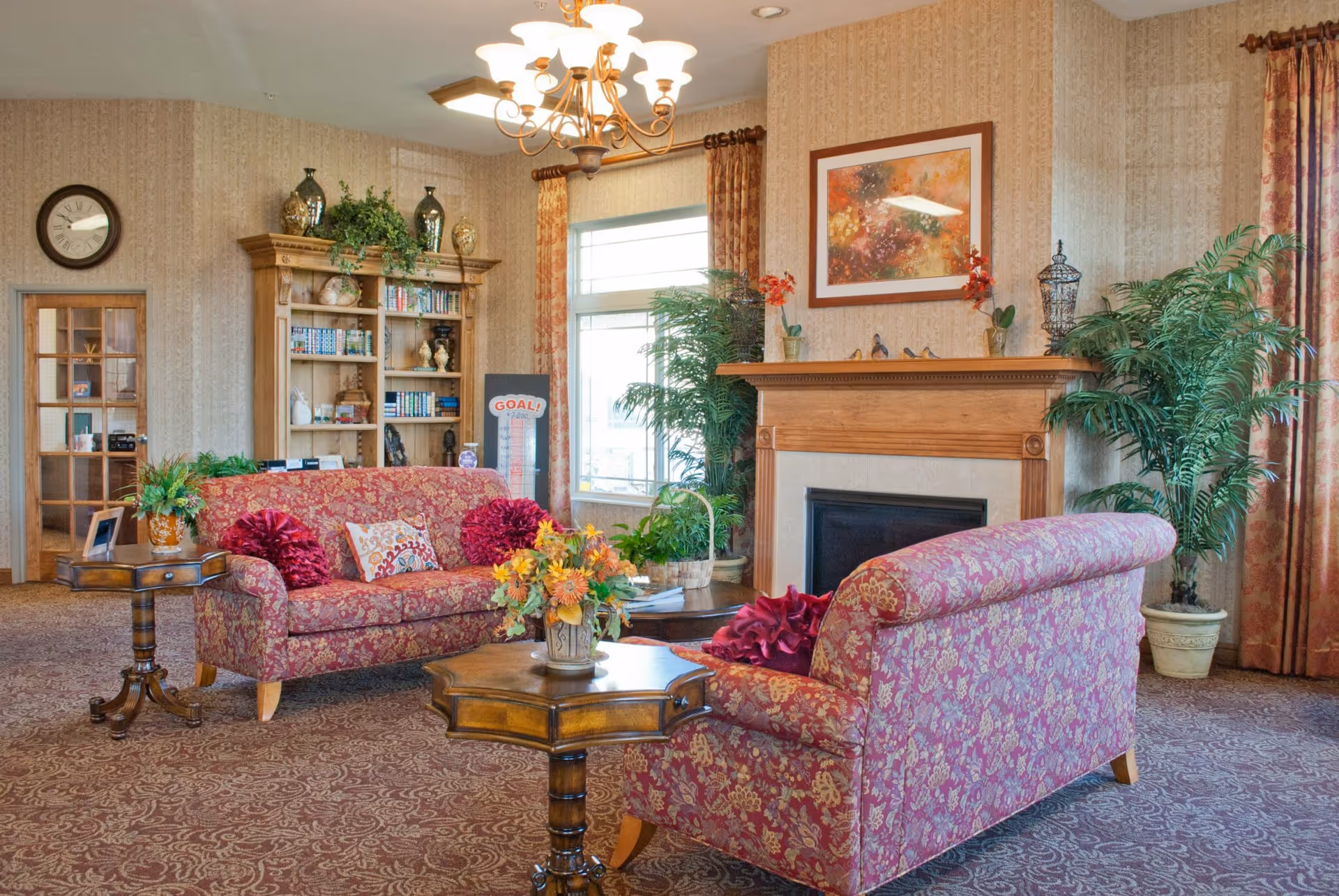 A cozy living room area in a senior living facility with two patterned red sofas, a wooden coffee table with a flower arrangement, a side table with a plant, a wooden bookshelf filled with books and decorative items, a fireplace with a wooden mantel decorated with small plants and bird figurines, a large window with floral curtains, and a wall clock.