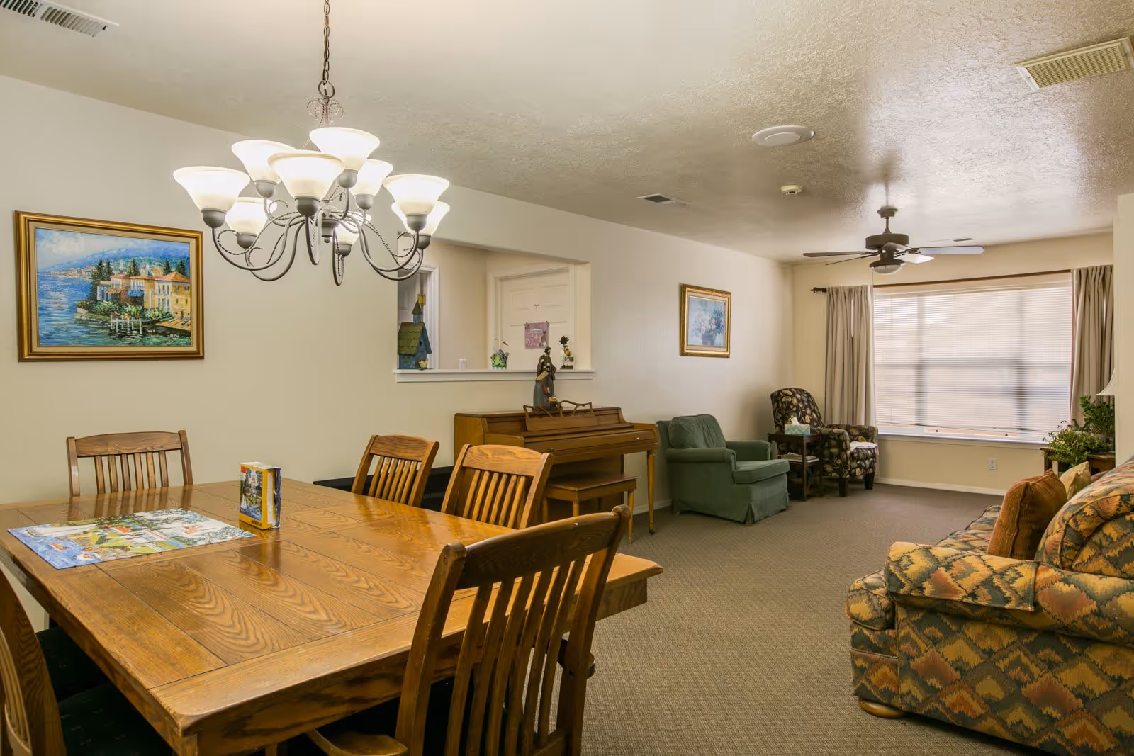 A combined dining and living room area in a senior living facility. The dining area features a wooden table with six chairs and a chandelier overhead. The living area includes a patterned sofa, two armchairs, a small side table, a piano, and framed paintings on the walls. A large window with curtains allows natural light into the room.