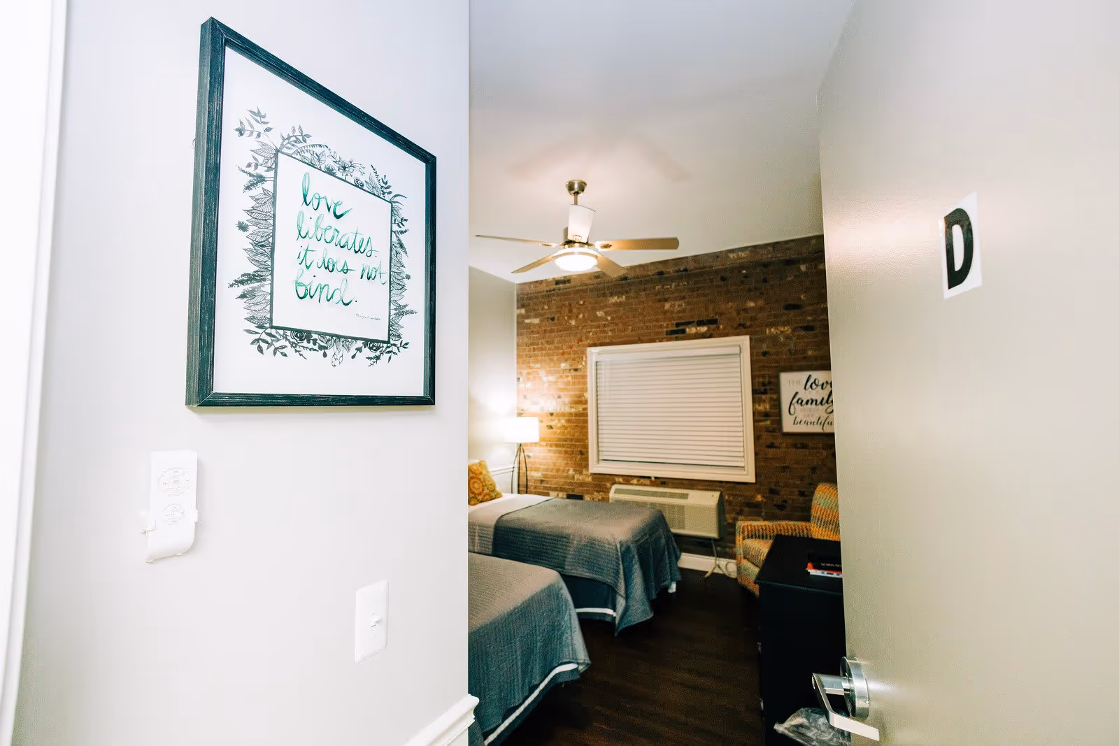 View into a bedroom with two twin beds covered in gray bedspreads, a ceiling fan with light, a brick accent wall with a window covered by blinds, a floor lamp, an armchair, and framed wall art. The door to the room is open with a letter D on it, and there is a framed quote on the adjacent wall that reads 'love liberates it was not bind.'