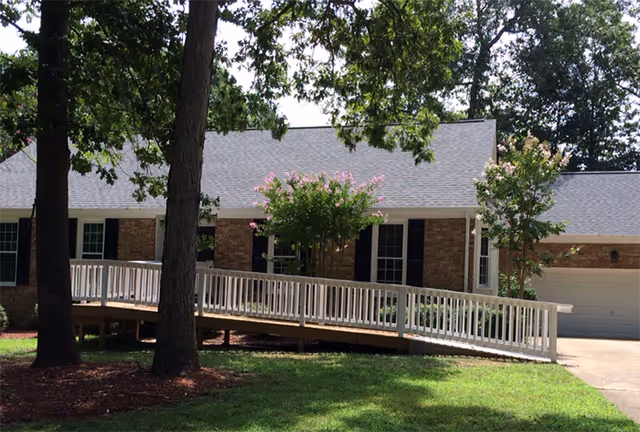 Exterior view of a single-story brick building with a gray roof, white railing ramp, and surrounded by trees and green grass under a clear sky.