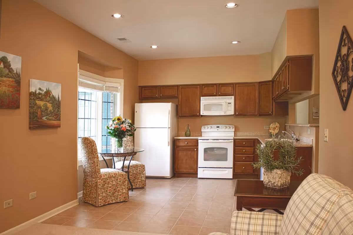 A cozy kitchen and dining area in an independent senior living facility. The kitchen features wooden cabinets, a white refrigerator, stove, and microwave. There is a small round glass table with two floral upholstered chairs near a window. A vase with flowers is on the table, and a potted plant is on a wooden side table next to a plaid armchair. The walls are painted a warm beige color, and there are two landscape paintings on one wall.