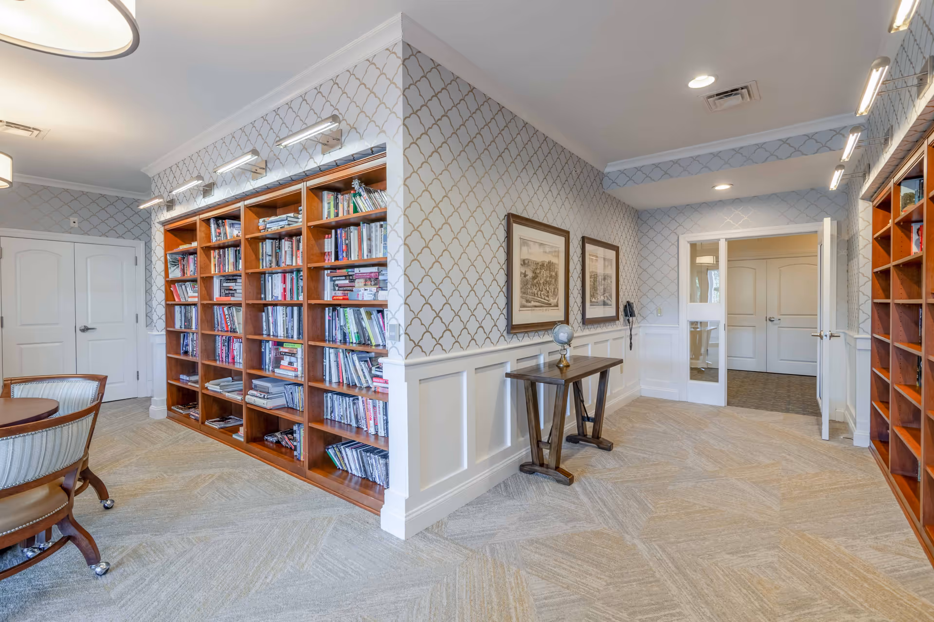 Well-lit interior reading/library area with built-in wooden bookshelves, a small table and chairs, patterned wallpaper, and double doors.