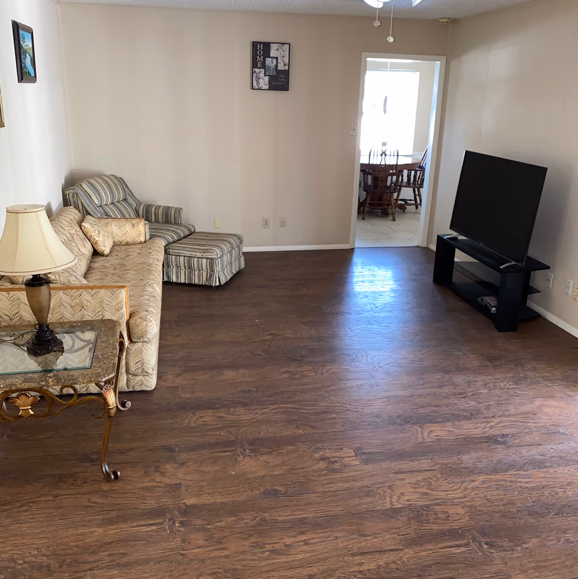 Spacious living room with a sofa and chaise, TV on a stand, side table with lamp, and wood flooring leading to a dining area.