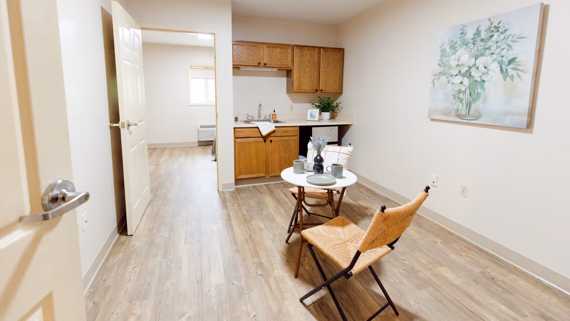 Small apartment living area with a kitchenette, a round table and two chairs on wood flooring and floral wall art.