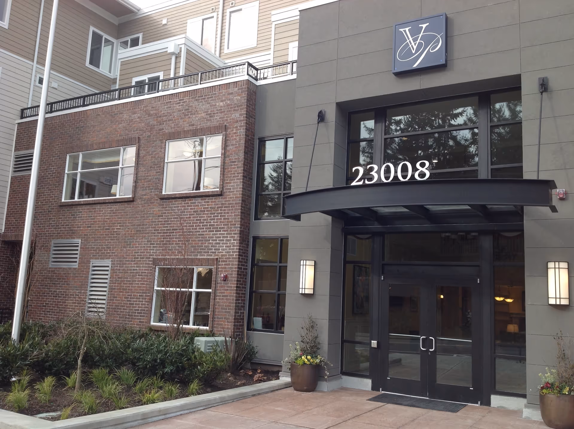 Exterior entrance of a senior living facility with a modern design featuring a combination of brick and gray paneling. The entrance has double glass doors under a black awning with the number 23008 displayed above. There are two wall-mounted lights on either side of the door and potted plants near the entrance. The building has multiple windows and some landscaping with shrubs and small trees in front.