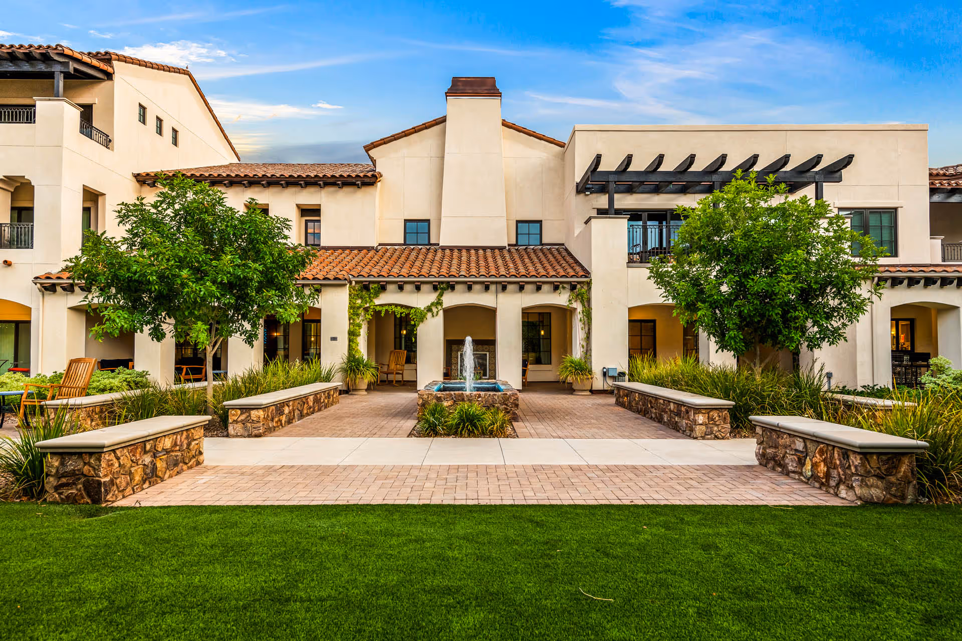 Exterior view of The Hacienda at the Canyon showing a beige stucco building with a red tile roof, a central water fountain, green trees, and well-maintained landscaping with grass and stone benches.