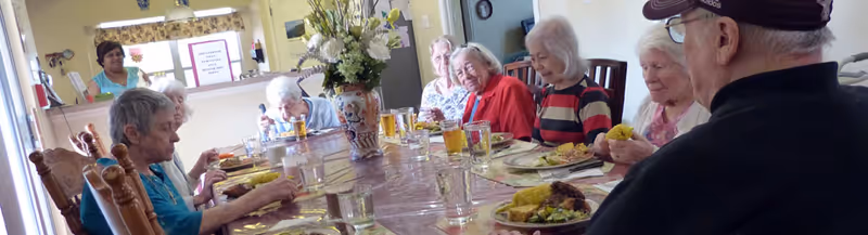 A group of elderly people sitting around a long dining table in a well-lit room, eating a meal together. The table is set with plates of food, glasses of water and juice, and a large floral centerpiece. The atmosphere appears communal and friendly.