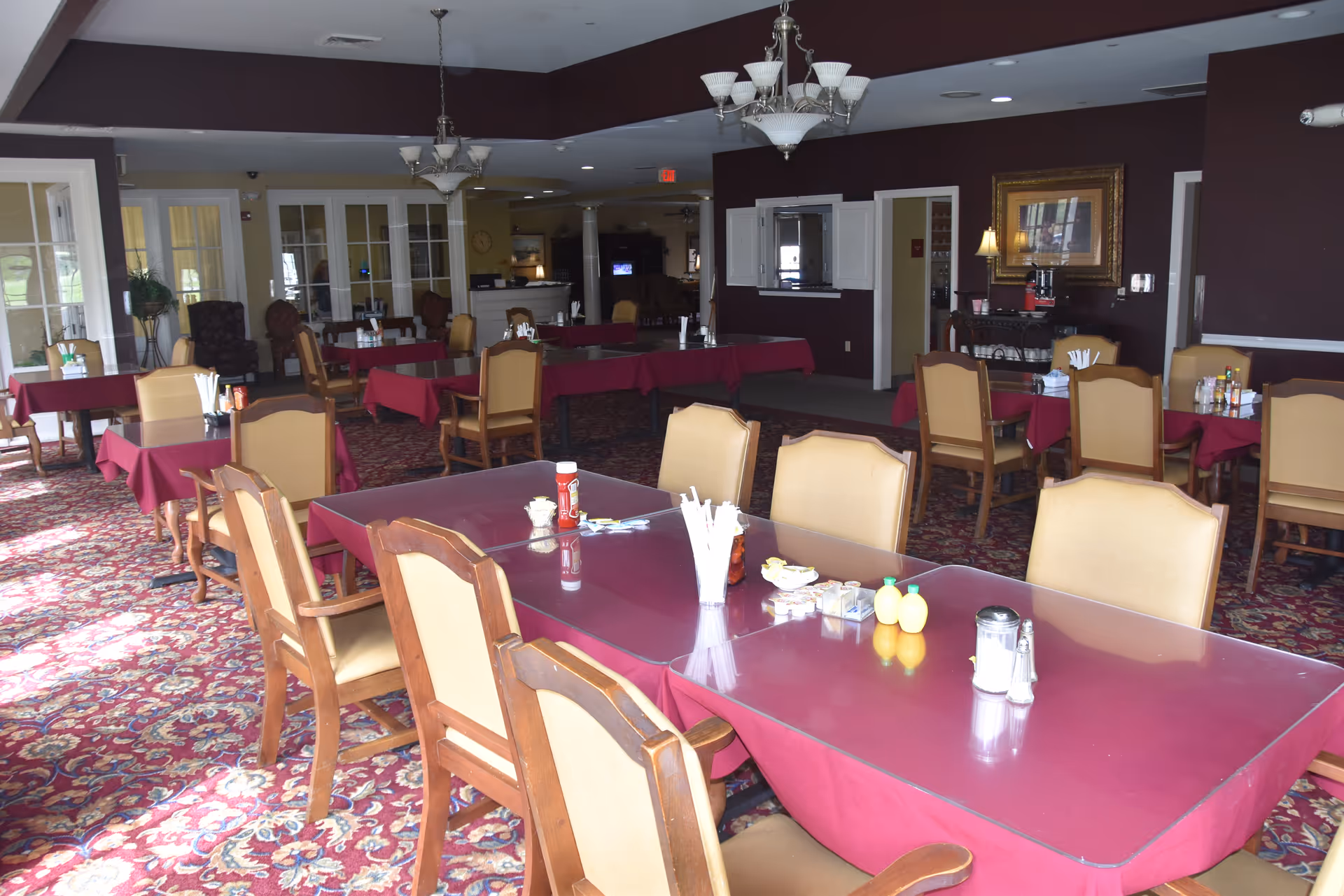 Dining room with multiple tables covered in red tablecloths, wooden chairs, chandeliers, and a patterned carpet in an assisted living facility.