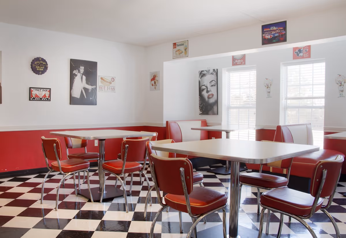 A retro-style dining room with red and white booths and chairs, black and white checkered floor, and vintage wall art including images of Elvis Presley and Marilyn Monroe. The room has large windows with white blinds allowing natural light to enter.