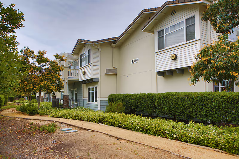 Exterior view of a two-story residential building with beige and light blue siding, surrounded by green shrubs and trees along a curved dirt pathway under a cloudy sky.
