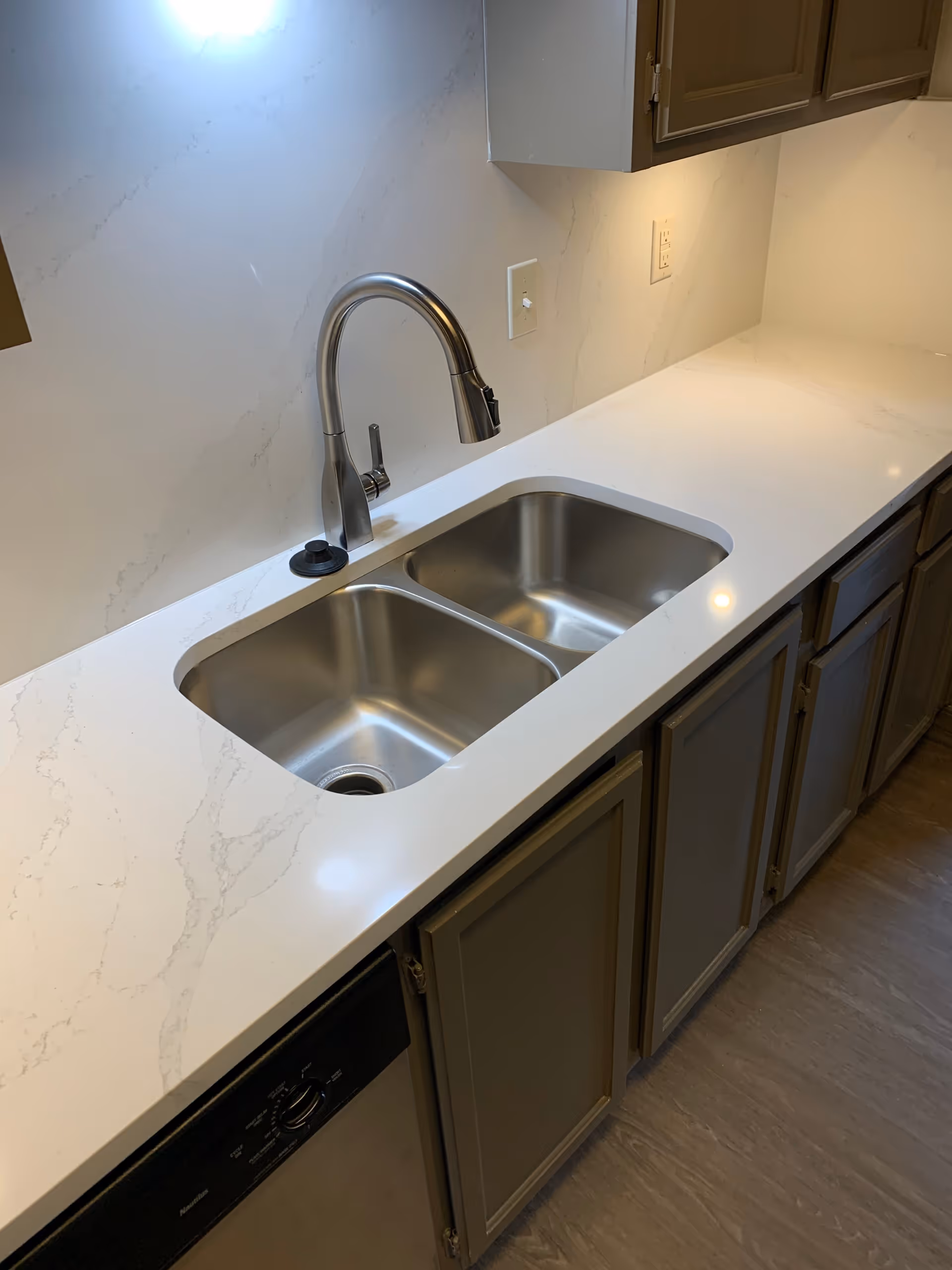 A modern kitchen countertop with a double stainless steel sink and a sleek, curved faucet. The countertop is white with subtle gray veining, and there are gray cabinets below and above the counter. The floor is a light wood tone, and there are electrical outlets on the wall above the counter.