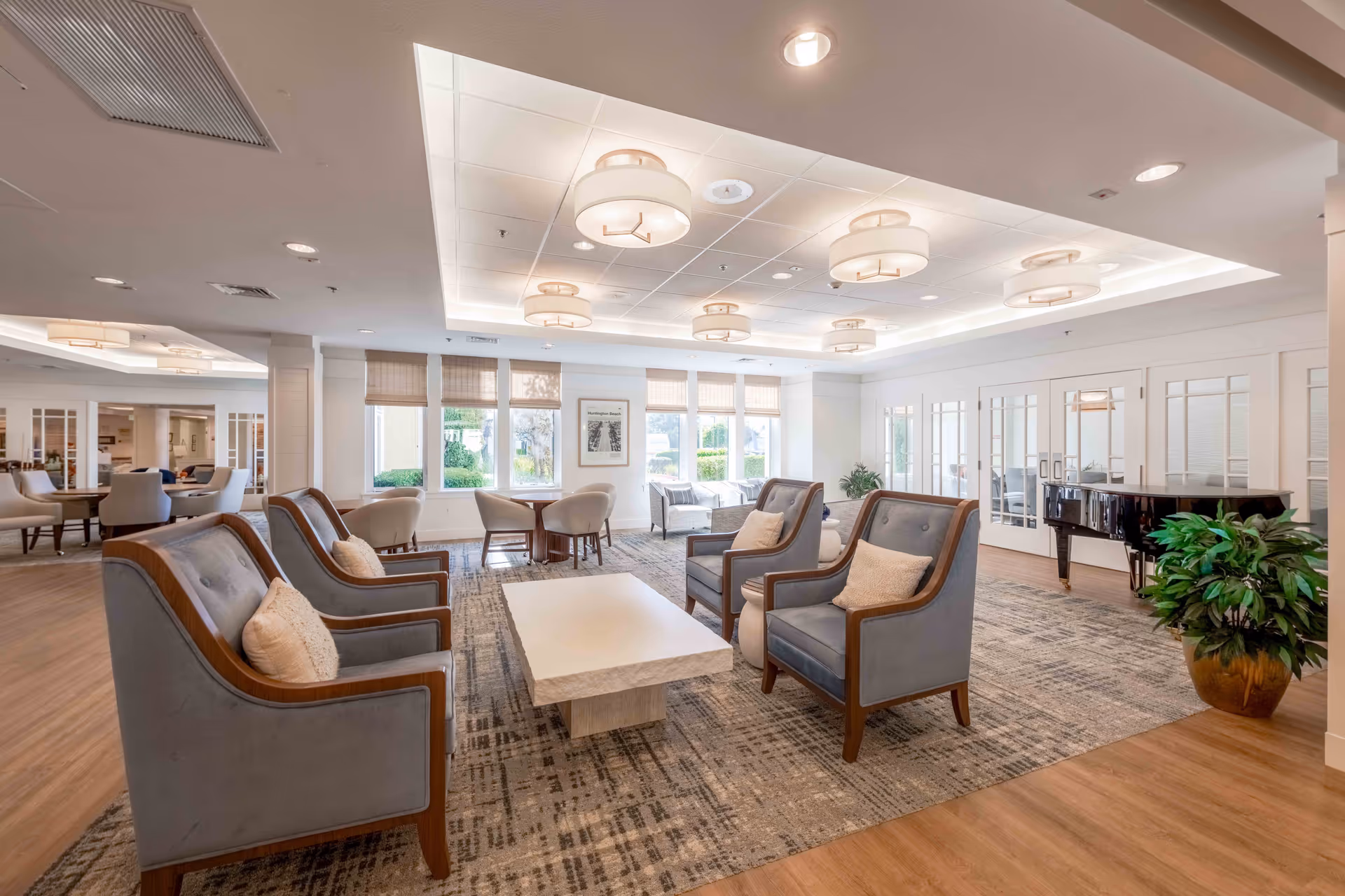 A bright and spacious senior living facility common area with multiple gray armchairs arranged around a rectangular white coffee table on a patterned rug. The room features large windows with beige blinds, modern ceiling lights, a black grand piano, and potted plants. There are additional seating areas with tables and chairs visible in the background.
