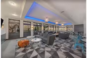 Modern lounge area with gray sofas, white round tables, an orange ottoman, and blue chairs. The ceiling features blue accent lighting and hanging spherical light fixtures. Large windows line the walls, allowing natural light to fill the space. The floor is covered with a geometric patterned carpet.