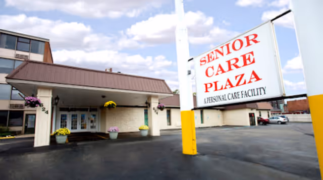 Exterior view of Senior Care Plaza, a personal care facility, showing the entrance with a covered drop-off area, flower pots with colorful flowers, and a large sign with the facility's name. The sky is partly cloudy.