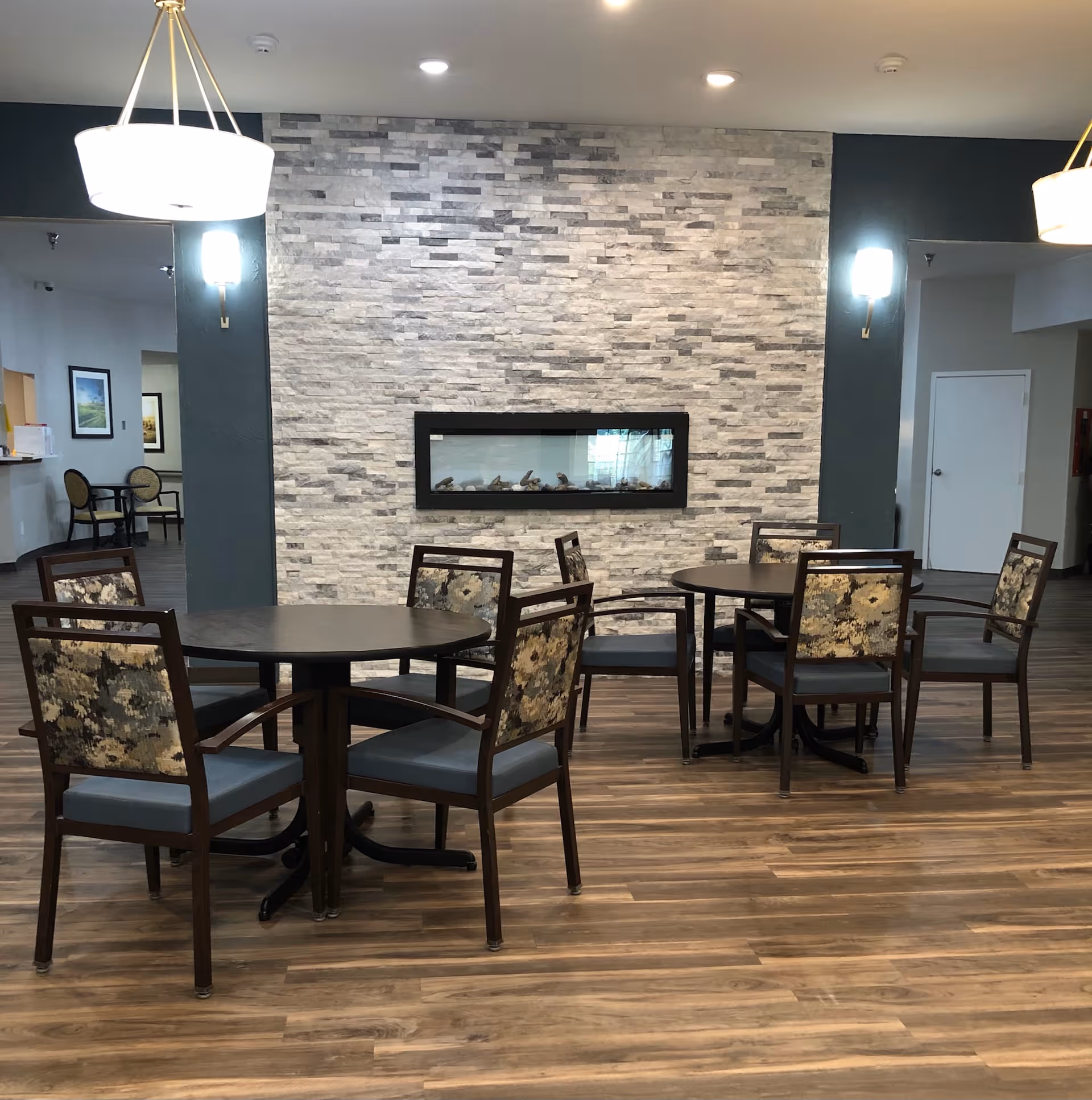 Communal dining area with round tables and upholstered chairs facing a stone accent wall with a horizontal fireplace.