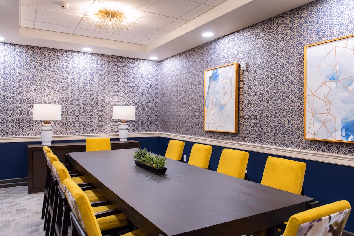 A modern conference or dining room with a long dark wooden table surrounded by yellow upholstered chairs. The walls have a blue and white geometric patterned wallpaper with two abstract paintings framed in gold. Two white table lamps sit on a sideboard against the wall, and a rectangular planter with green plants is centered on the table. The ceiling has recessed lighting and a decorative light fixture.
