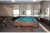 A room with a pool table in the center, surrounded by folding chairs along the left wall. Large windows with blinds cover the walls, allowing natural light to fill the space. A potted plant is placed in the corner near the windows.