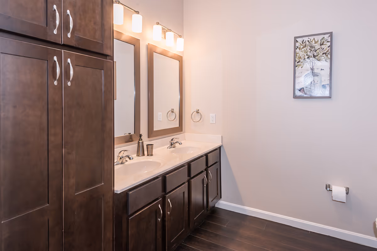 A bathroom interior featuring a double sink vanity with dark wood cabinets and two rectangular mirrors above each sink. Above the mirrors are light fixtures with four lights. On the right wall, there is a framed artwork of a vase with leaves and a toilet paper holder with a roll of toilet paper. The floor is dark wood, and the walls are painted light beige.