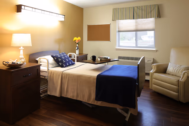 A neatly arranged room in a senior living facility with a single bed featuring beige bedding and a navy blue blanket. There is a navy blue pillow with white anchor patterns on the bed. Next to the bed is a wooden nightstand with a lamp and a basket. A small table with a black mug and books is positioned over the bed. The room has a window with a striped valance and a beige armchair with a pillow that says 'Home'. The walls are painted in warm tones and the floor is wooden.