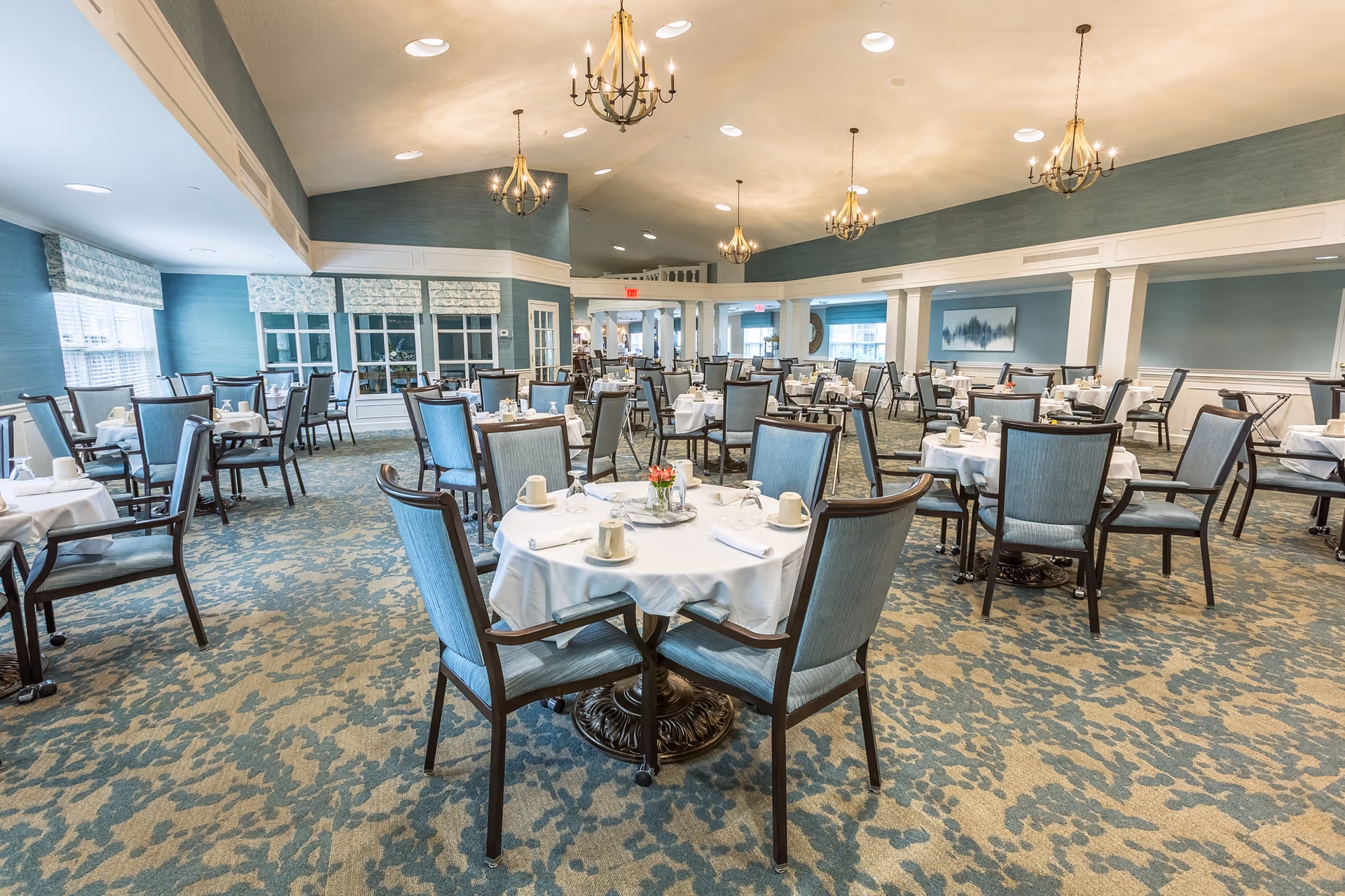 Spacious dining room with round tables set with white tablecloths, blue upholstered chairs, and chandeliers.