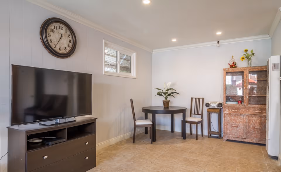 A cozy interior room with a large flat-screen TV on a dark wooden stand, a round table with two chairs, a potted plant on the table, a wooden cabinet with glass doors, a small stereo on a stand, and a large wall clock above the TV. The room has light-colored walls and tiled flooring.
