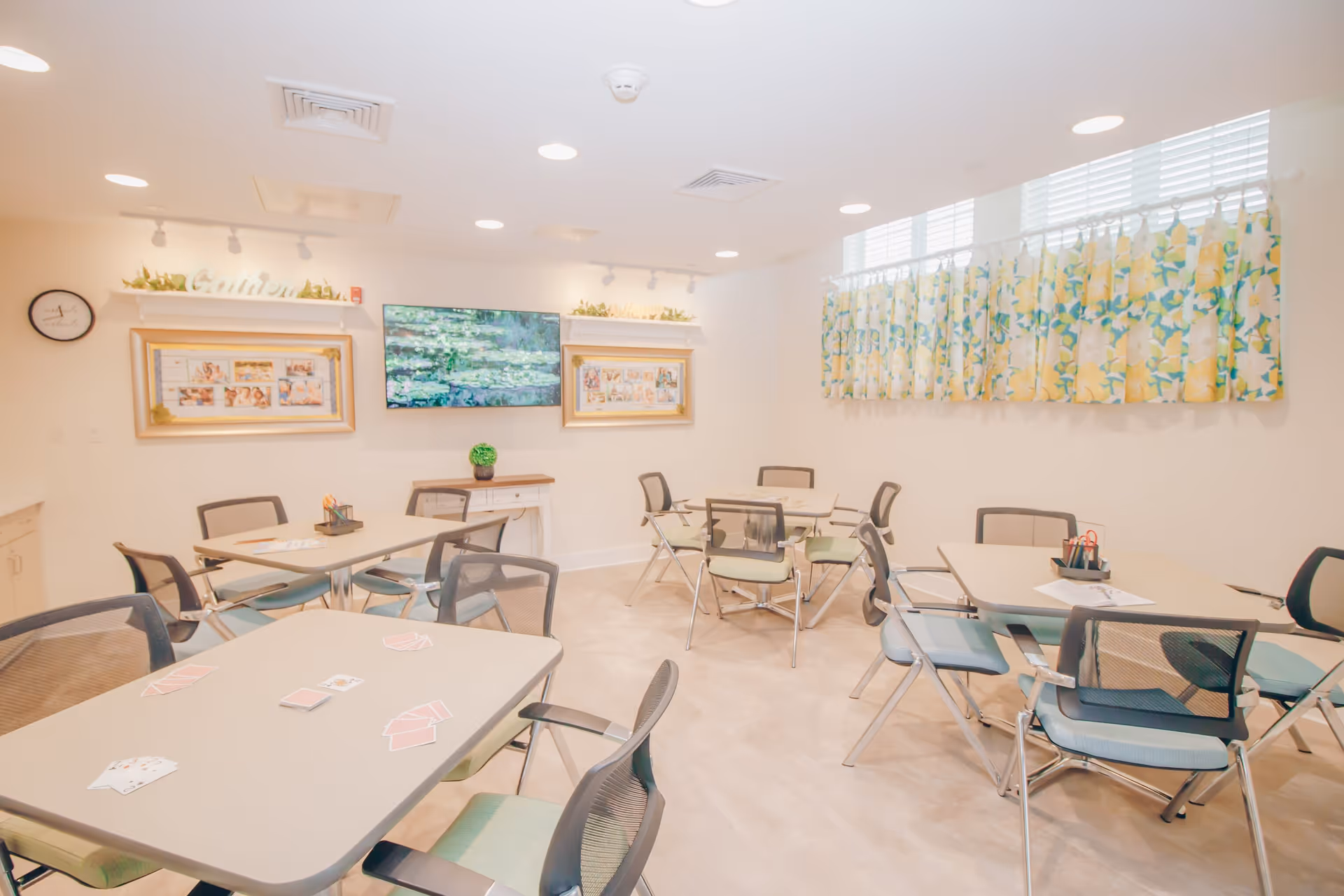 A bright and clean activity room with several tables and chairs arranged for group activities. Playing cards are spread out on one table. The room has light-colored walls, a wall-mounted TV displaying a nature scene, framed pictures, and a window with floral curtains. The ceiling has recessed lighting and air vents.