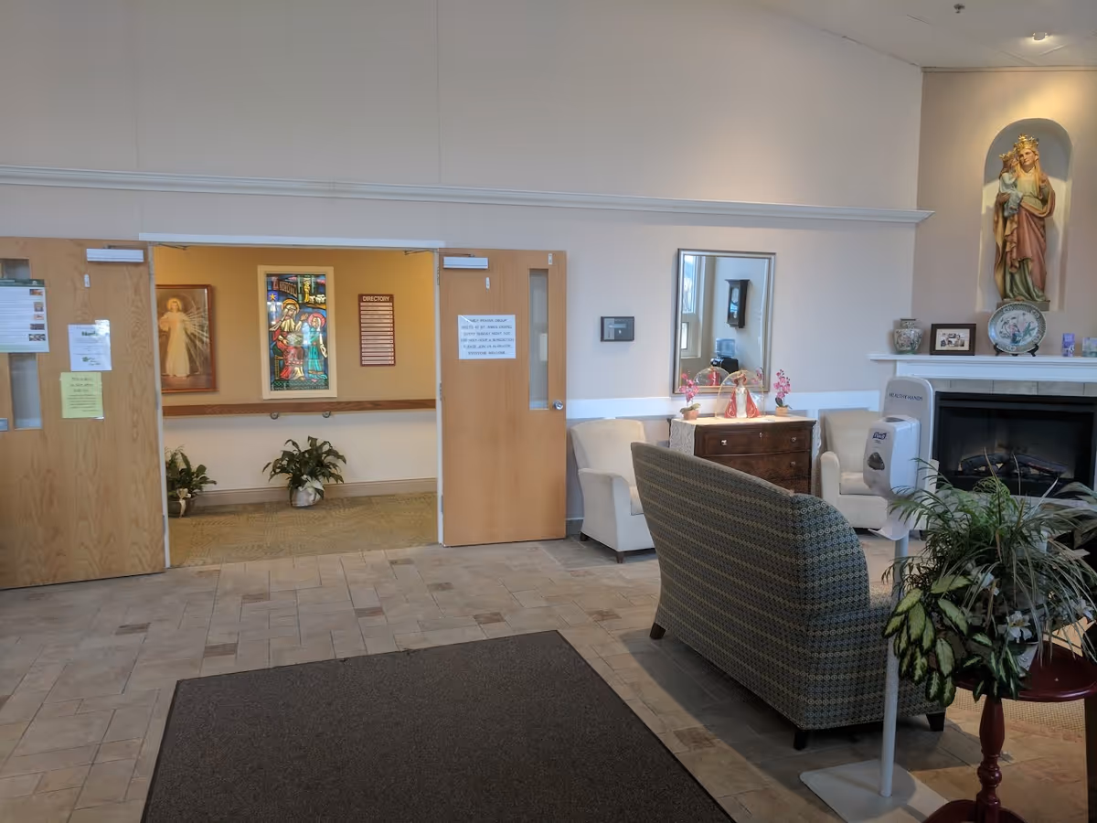 Interior view of a common area in St. Ann's Home featuring a seating arrangement with two armchairs and a patterned loveseat around a small wooden dresser with decorative items. A statue of a religious figure is displayed in a niche above a fireplace. There are double wooden doors leading to a hallway with religious artwork and plants. A hand sanitizer dispenser is visible near the seating area.