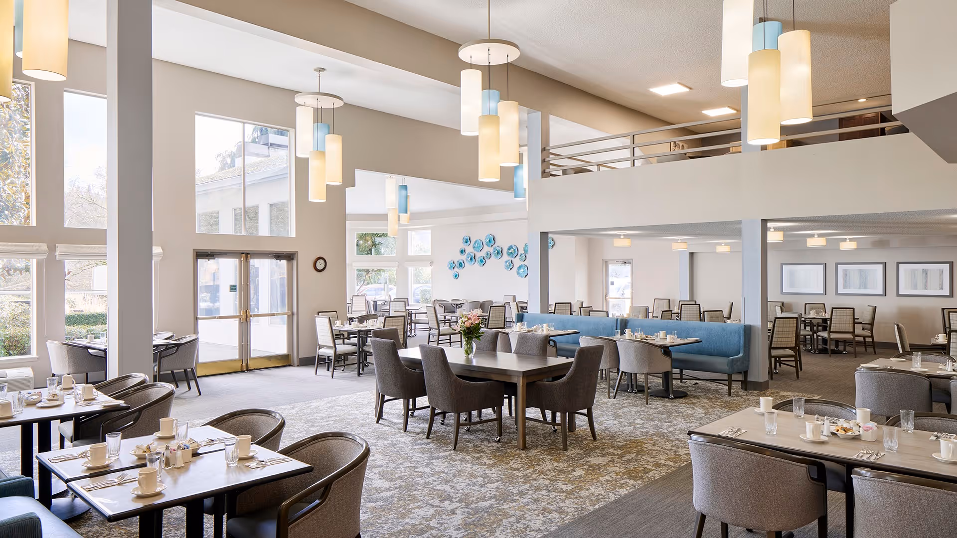 A spacious and well-lit dining room in a senior living facility with multiple tables and chairs arranged for meals. The room features large windows allowing natural light, modern pendant lighting, and a mix of seating including cushioned chairs and booth-style benches. Tables are set with cups, glasses, and utensils, ready for dining.