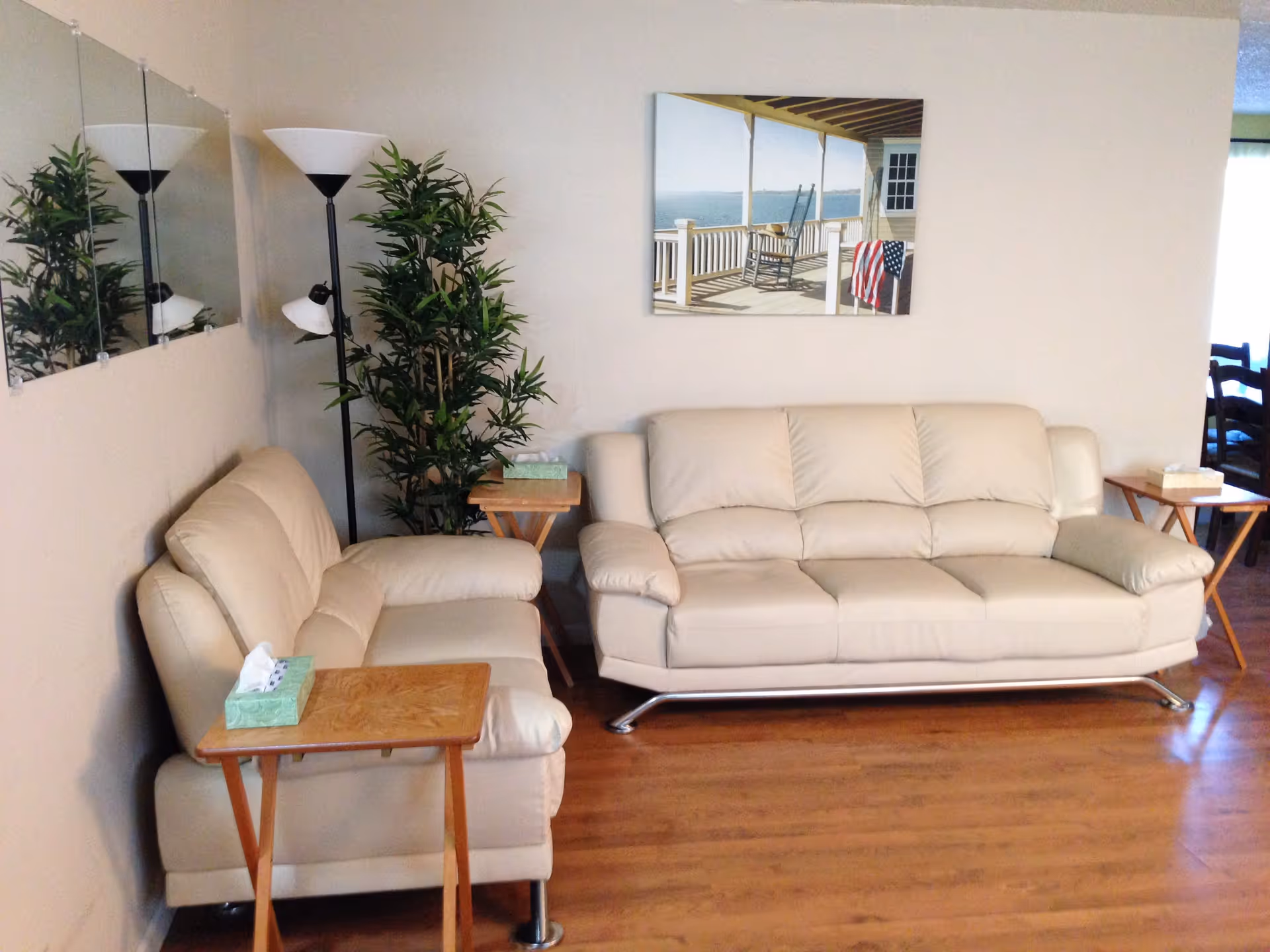 A living room with two beige leather sofas facing each other, each accompanied by a wooden side table with a tissue box on top. There is a tall floor lamp and a green potted plant in the corner. A painting of a porch with a rocking chair and an American flag hangs on the wall above one sofa. The floor is wooden, and a mirror is mounted on the adjacent wall.