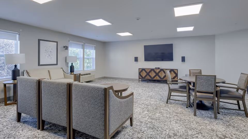 A spacious living room with gray patterned carpet, a set of upholstered chairs and a sofa arranged around a round coffee table, two side tables with lamps, a round dining table with four chairs, and a flat-screen TV mounted on the wall above a wooden cabinet.