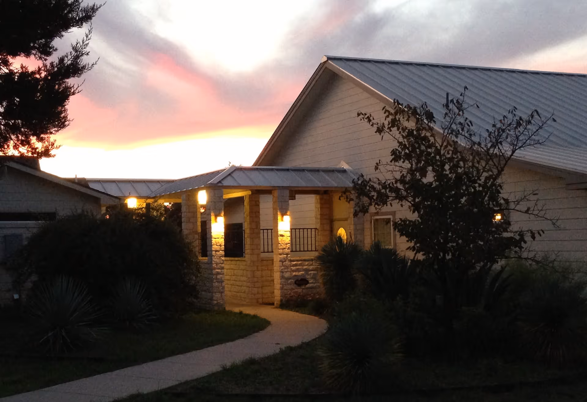 Exterior view of a building at sunset with a covered entrance illuminated by warm lights, surrounded by bushes and trees.