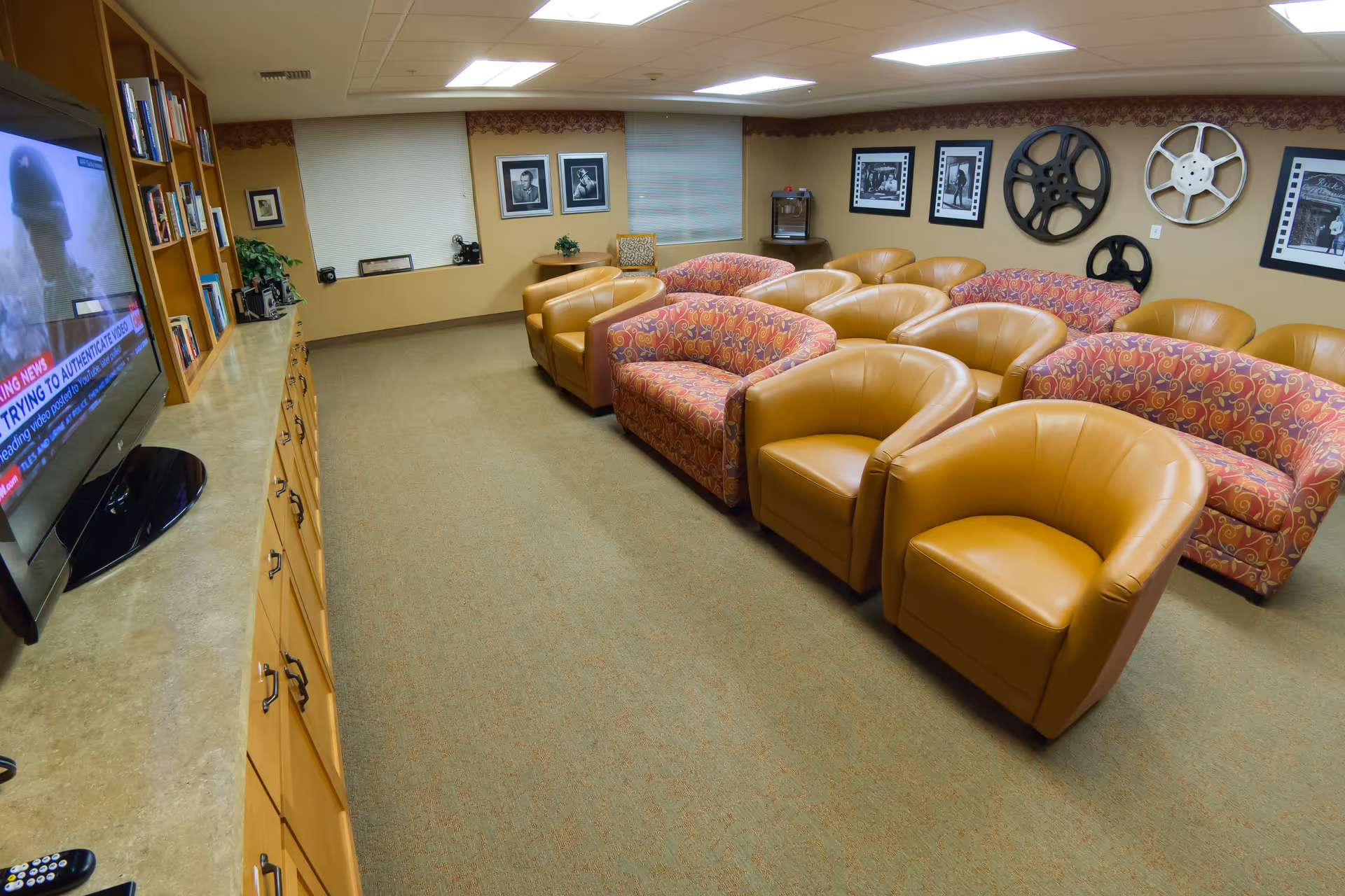 A cozy media room with multiple rows of comfortable armchairs and sofas facing a large flat-screen TV mounted on a wooden cabinet. The walls are decorated with framed black and white photos and large vintage film reels. The room has beige carpet and warm yellow walls with a window covered by blinds.