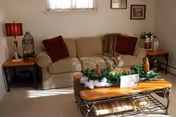 A cozy living room with a beige sofa adorned with two red cushions and a brown throw blanket. There are two wooden side tables on either side of the sofa, one with a red lamp and decorative items, and the other with a vase of flowers. A wooden coffee table with metal legs is in front of the sofa, decorated with a festive green garland and holiday ornaments. The room has light-colored walls and carpet, with framed pictures hanging on the wall.