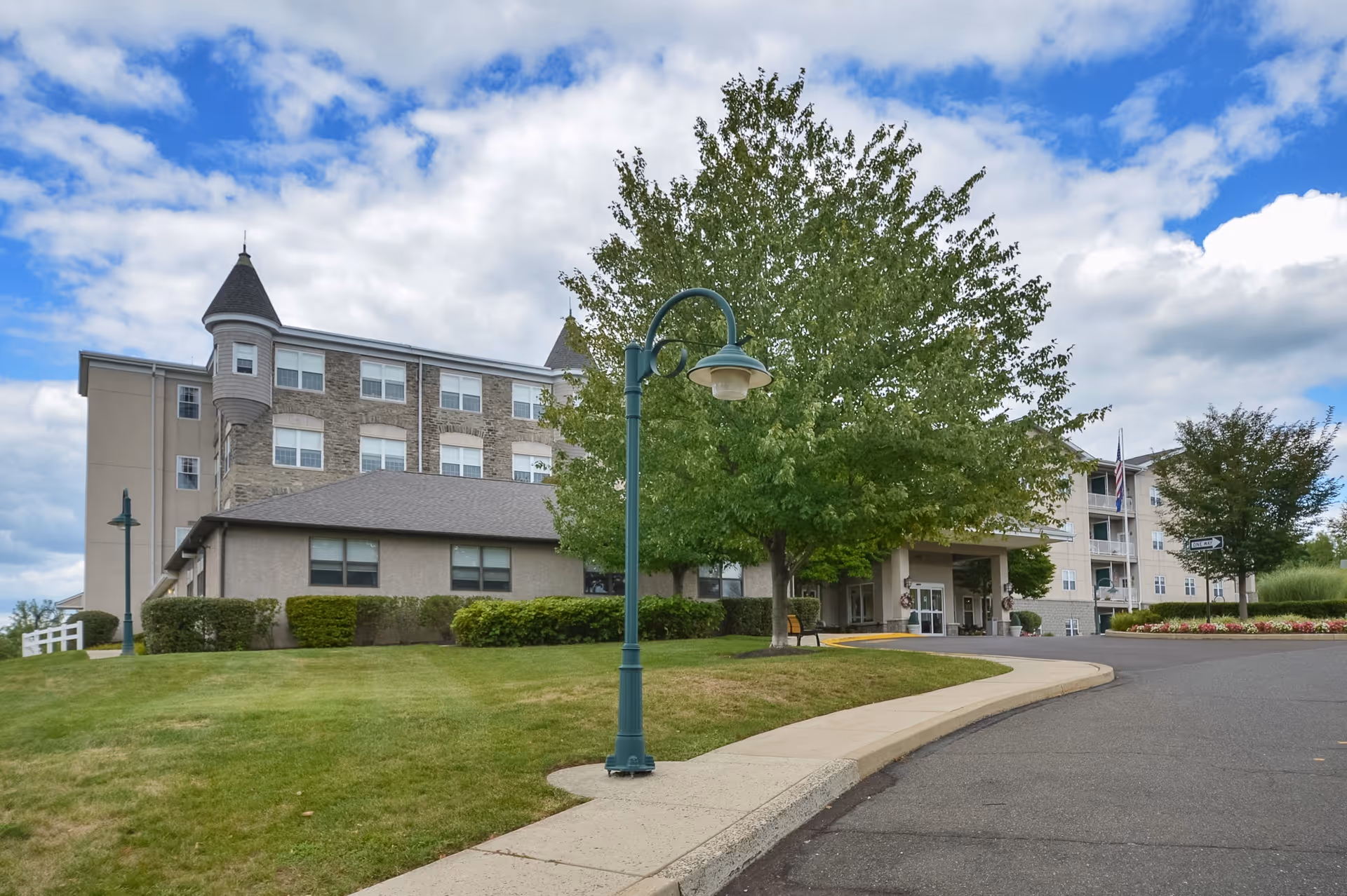 Exterior view of a multi-story senior living facility building with a turret-like architectural feature, surrounded by green lawns, trees, and a curved driveway under a partly cloudy sky.