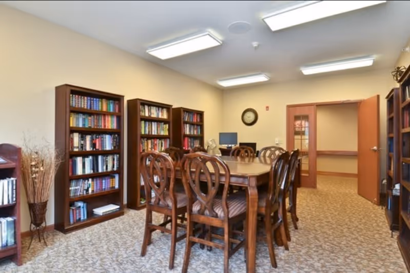 A well-lit room with beige walls and carpeted floor featuring a large wooden table surrounded by eight matching wooden chairs. The room has multiple wooden bookshelves filled with books along the walls. A clock is mounted on the far wall above a computer workstation with a monitor and keyboard. The room has fluorescent ceiling lights and an open door leading to a hallway.