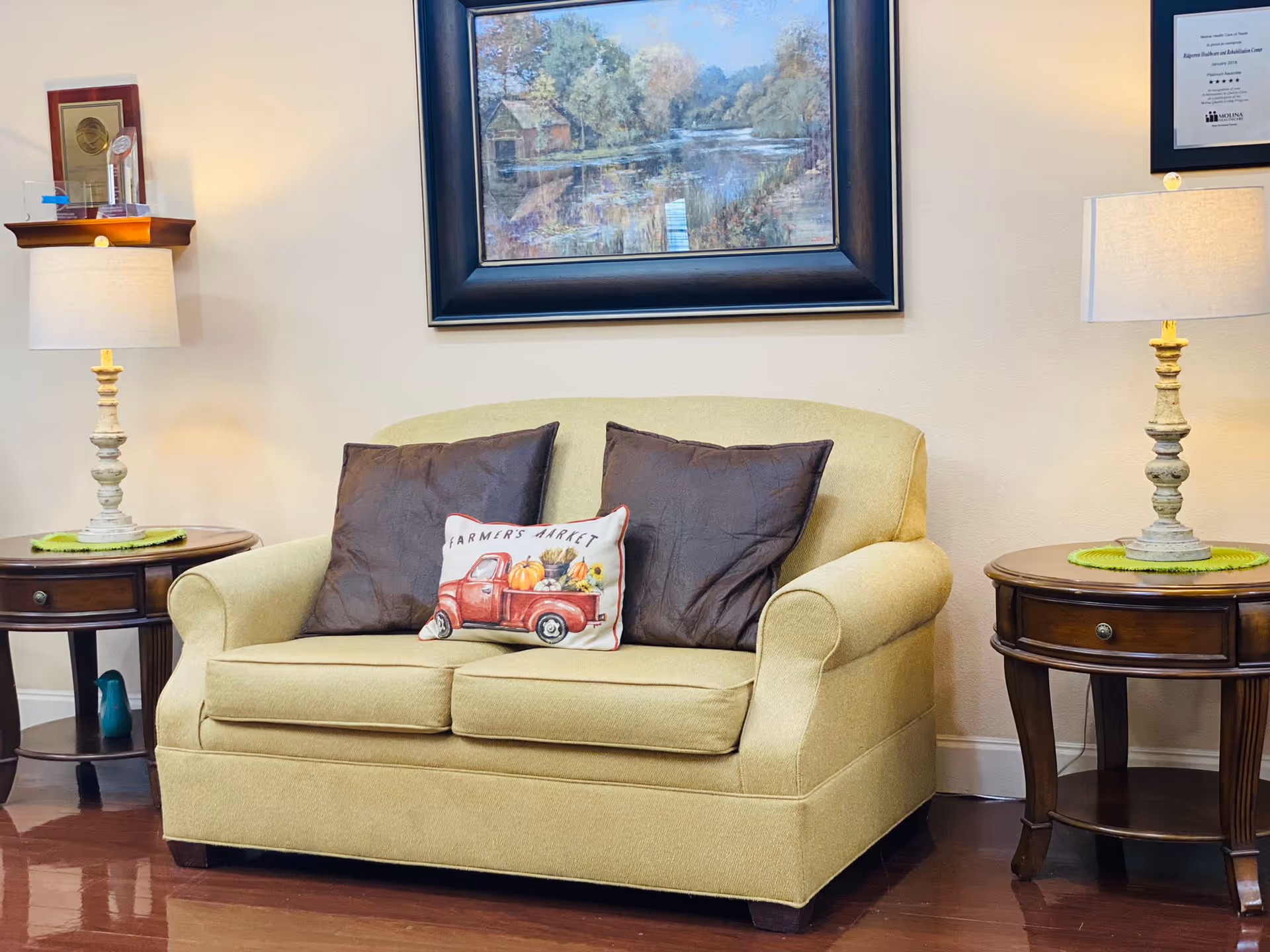 A cozy seating area with a light green loveseat adorned with two dark brown pillows and a decorative pillow featuring a red truck and the words 'Farmers Market'. On either side of the loveseat are wooden side tables with drawers, each topped with a lamp. Above the loveseat hangs a framed painting of a serene lakeside scene with trees and a small dock.